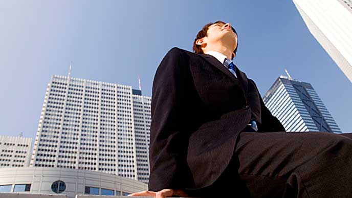 企業ビルが立ち並ぶ街で空を見上げるスーツ姿の男性